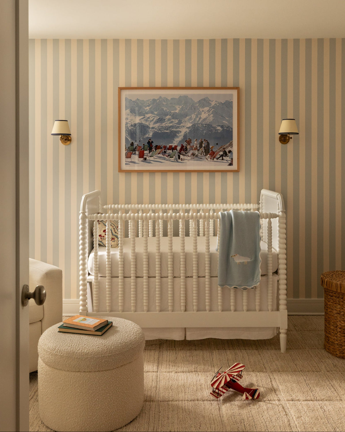 A styled nursery interior featuring a white wooden crib with a light blue blanket, striped wallpaper, a round beige ottoman, and a framed mountain landscape painting on the wall.