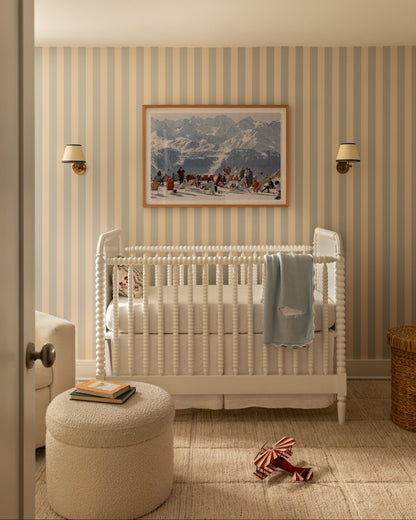 A styled nursery interior featuring a white wooden crib with a light blue blanket, striped wallpaper, a round beige ottoman, and a framed mountain landscape painting on the wall.